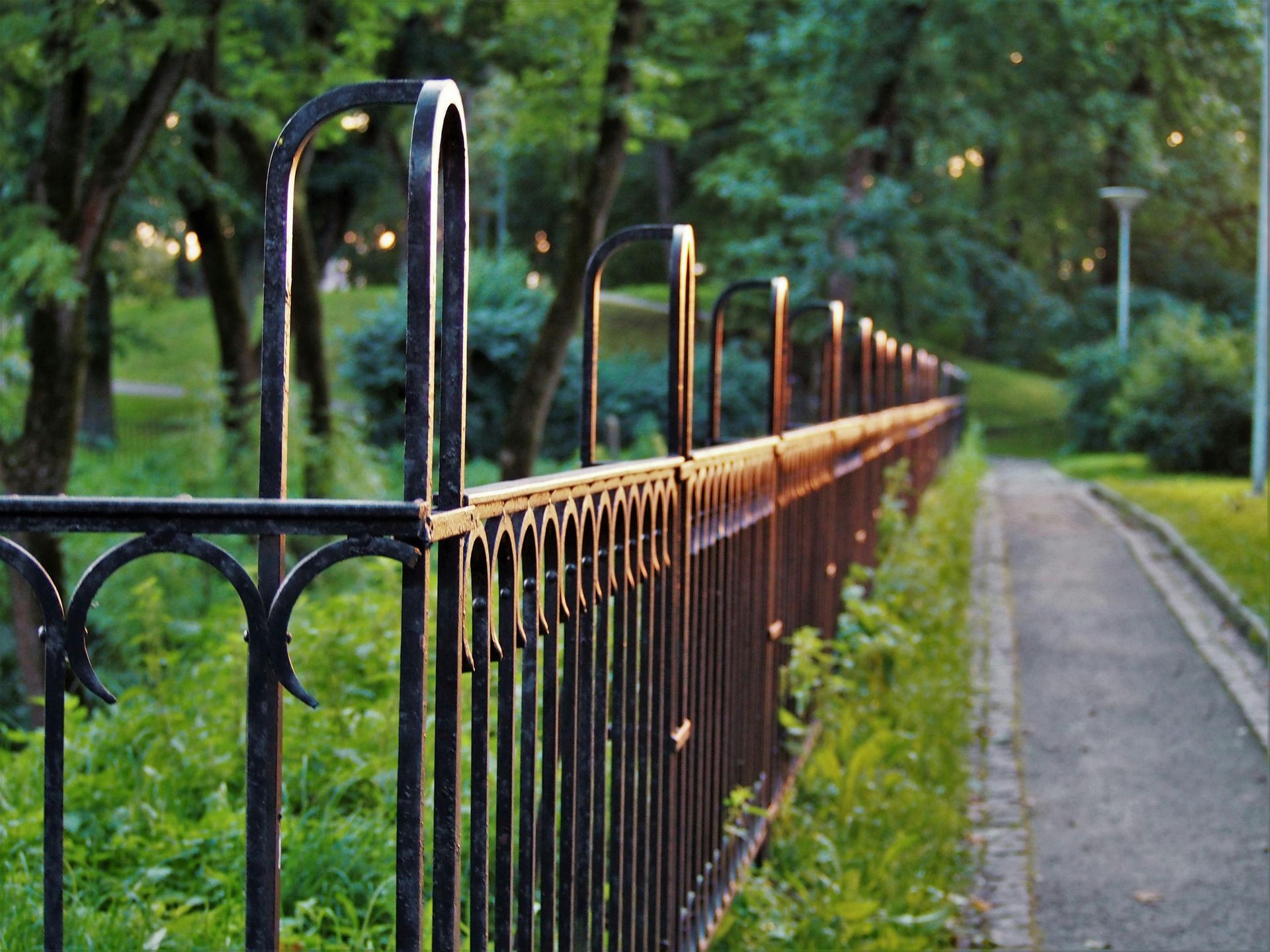 Black wrought-iron fence alongside a paved path in a green park with trees.